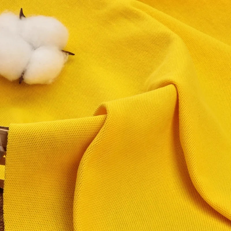 Un Tissu piqué uni au mètre jaune vif de Longan Craft France est drapé et plié, avec du coton brut sur un coin - parfait pour les polos d'été DIY ou les chemises à manches courtes en coton-polyester.