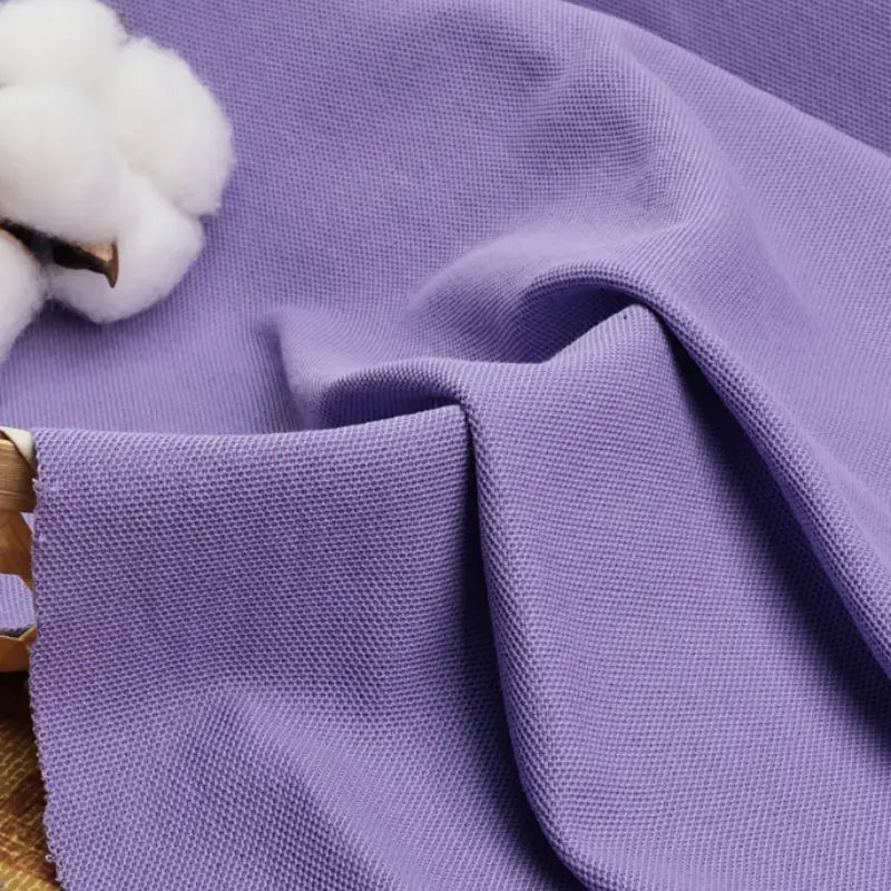 Un échantillon de Tissu piqué uni au mètre de Longan Craft France, un mélange de coton et de polyester idéal pour les polos et les chemises d'été, est drapé sur une surface à côté d'une balle de coton, mettant en valeur son tissage texturé et sa douceur.