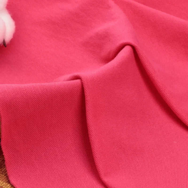 Un gros plan du tissu piqué uni au mètre rose vif de Longan Craft France montre son tissage doux et texturé en coton-polyester avec des plis visibles. Une partie d'un objet blanc apparaît dans le coin supérieur gauche. Parfait pour les chemises et polos d'été DIY.