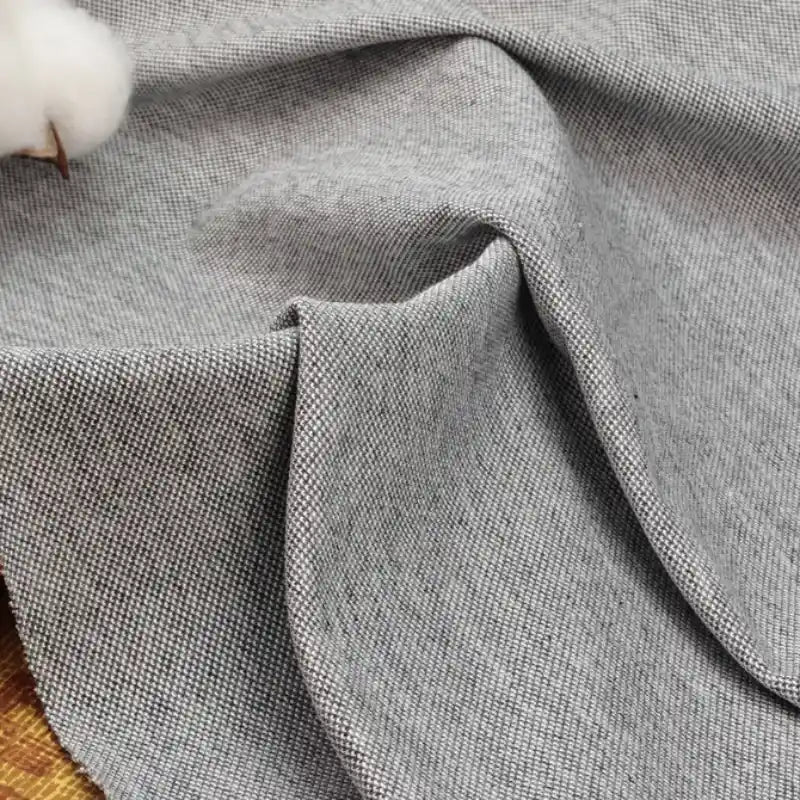 Un gros plan du piqué coton-polyester gris clair de Longan Craft France montre des plis doux et un tissage visible, avec une balle de coton mettant en valeur ce tissu classique pour polos et chemises d'été, vendu au mètre pour projets DIY.