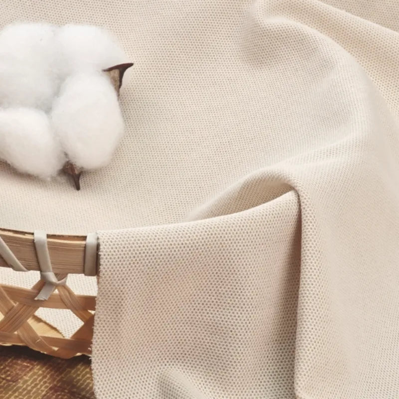 Gros plan sur le tissu piqué uni au mètre beige de Longan Craft France, un tissu coton-polyester pour polos et chemises d'été DIY, drapé sur un panier avec du coton brut reposant sur sa surface texturée.