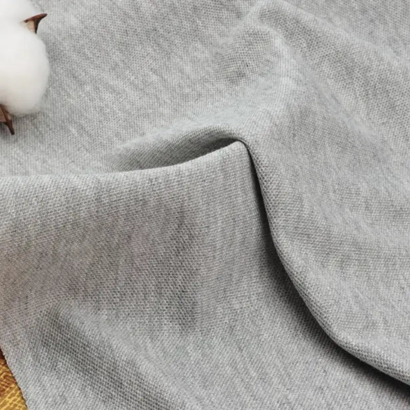 Un morceau de tissu piqué gris clair uni au mètre de Longan Craft France est présenté avec des plis visibles et une balle de coton en haut à gauche, mettant en évidence sa surface douce et texturée en coton-polyester pour les chemises d'été et les polos.