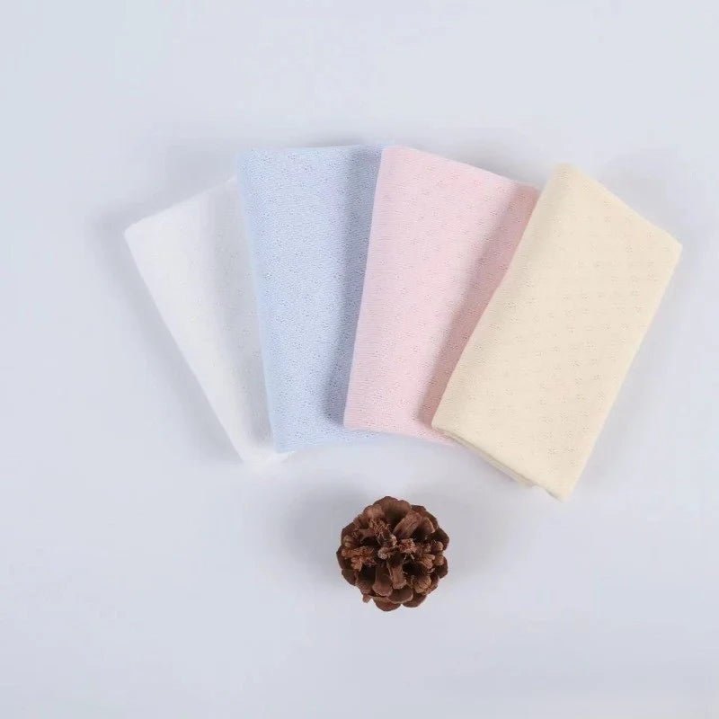 Quatre gants de toilette pliés en blanc, bleu clair, rose et crème, fabriqués en maille pointelle côtelée en coton biologique de Longan Craft France, sont disposés côte à côte sur une surface unie, avec une pomme de pin brune placée en dessous.