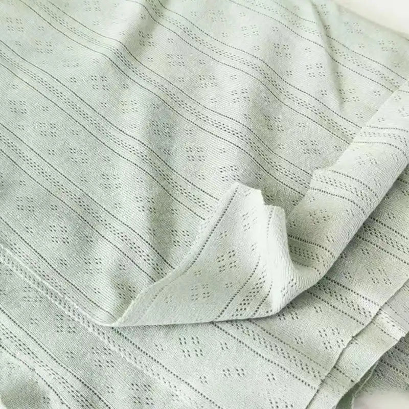 Gros plan sur la Maille pointelle côtelée unie au mètre de Longan Craft France, un tissu vert clair en coton biologique texturé avec de subtiles perforations en losange, idéal pour les vêtements bébé. Le tissu doux et respirant est légèrement plié et posé à plat.