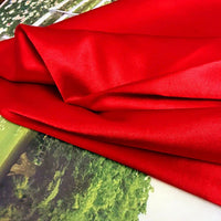 Un tissu satin polyester rouge brillant uni au mètre de Longan Craft France à la texture lisse est drapé sur une surface imprimée de verdure et d'une clôture, idéal comme tissu satin extensible pour des vêtements confortables et ajustés.