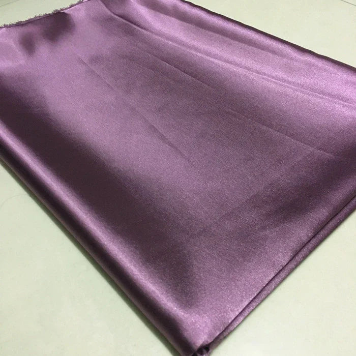 Un morceau de Longan Craft France Tissu satin polyester uni au mètre, violet clair avec une texture lisse et une brillance subtile, repose sur une surface pâle - idéal satin extensible pour vêtements ajustés et confortables.