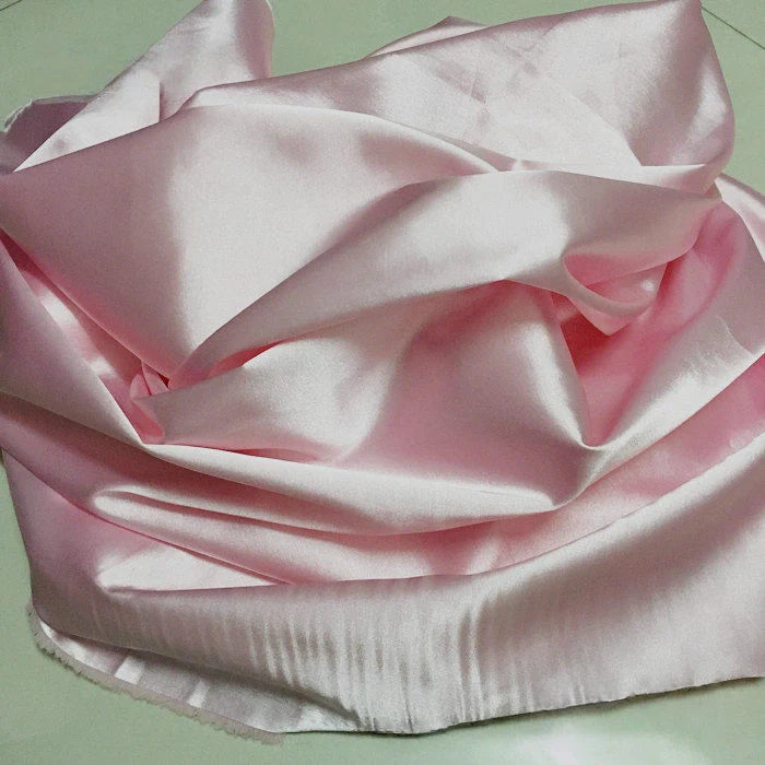 Un morceau de Tissu satin polyester uni au mètre de Longan Craft France en rose clair est rassemblé de manière lâche sur une surface plane et légère. Sa texture lisse et brillante et ses plis souples font de ce satin extensible un tissu idéal pour la confection de vêtements confortables et ajustés.
