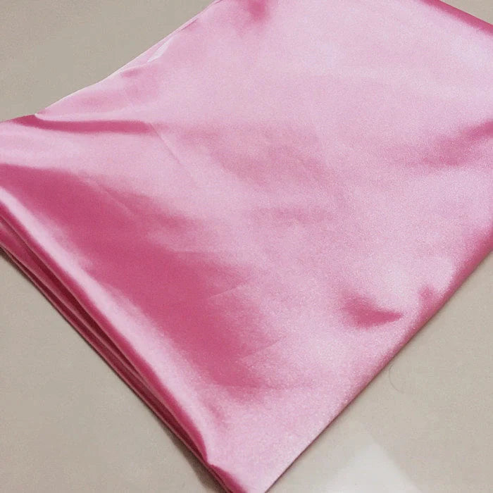 Un morceau plié de tissu satin polyester rose clair brillant Longan Craft France uni au mètre repose sur une surface neutre. Ce tissu satiné extensible est lisse, réfléchissant et idéal pour créer des vêtements ajustés et confortables.