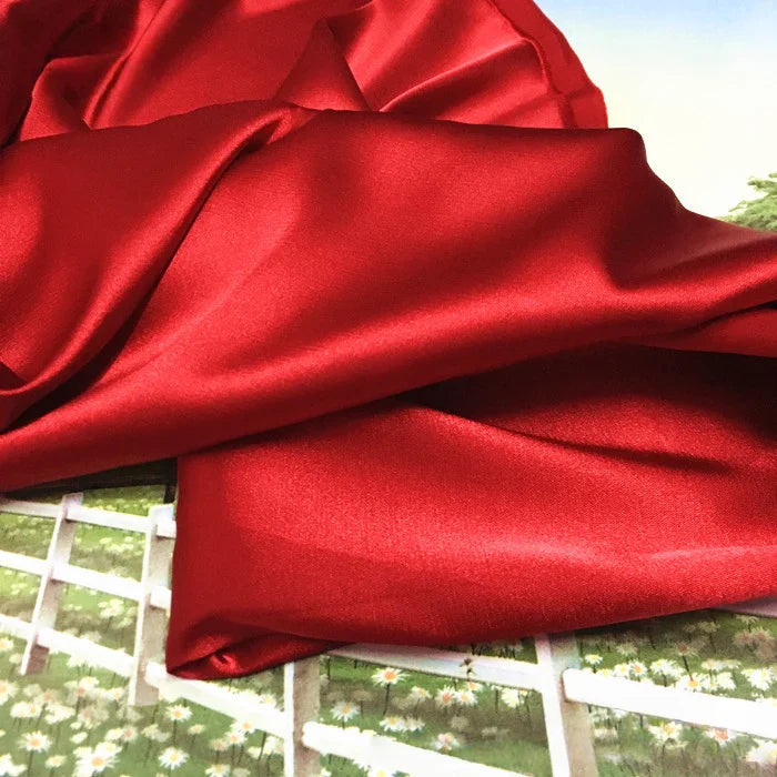 Le Tissu satin polyester uni au mètre de Longan Craft France, extensible et idéal pour vêtements ajustés, est présenté en rouge brillant sur une surface, avec une clôture blanche et un gazon fleuri en arrière-plan.