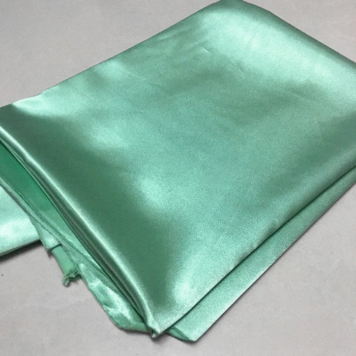 Une pièce de Longan Craft France Tissu satin polyester uni au mètre, vert clair avec une surface lisse et brillante, est présentée sur un fond uni - un satin parfait extensible pour créer des vêtements ajustés et confortables.