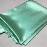 Une pièce de Longan Craft France Tissu satin polyester uni au mètre, vert clair avec une surface lisse et brillante, est présentée sur un fond uni - un satin parfait extensible pour créer des vêtements ajustés et confortables.