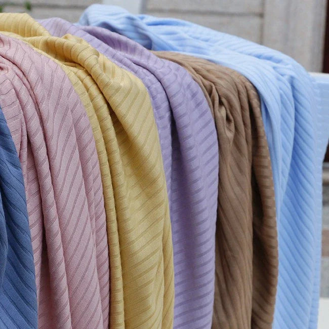 Plusieurs pièces de tissu côtelé uni en viscose au mètre de Longan Craft France de différentes couleurs sont suspendues côte à côte sur une tringle, sur fond de pierre. Ce textile extensible et confortable est idéal pour le bricolage de t-shirts.