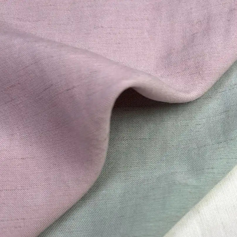 Un gros plan montre trois tissus superposés : mauve, gris-vert clair et blanc cassé. Le "Tissu uni en viscose lyocell" mauve de Longan Craft France est drapé sur le dessus, mettant en valeur sa texture lisse et respirante pour les hauts.