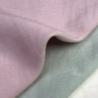 Un gros plan montre trois tissus superposés : mauve, gris-vert clair et blanc cassé. Le "Tissu uni en viscose lyocell" mauve de Longan Craft France est drapé sur le dessus, mettant en valeur sa texture lisse et respirante pour les hauts.