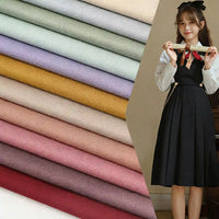 Une jeune femme en robe noire et chemise blanche se tient à côté de Longan Craft France Tissu gabardine uni au mètre soigneusement empilés dans des tons pastel et terreux - bleu clair, lilas, jaune moutarde, rose et rouge vin.