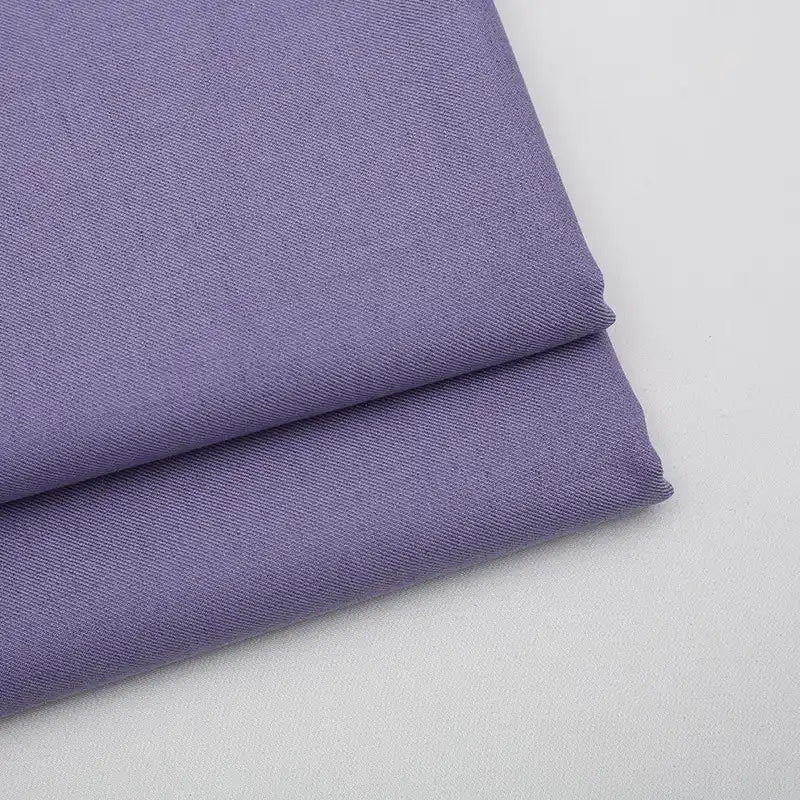 Un gros plan montre deux pièces soigneusement pliées de Tissu gabardine uni au mètre de Longan Craft France en violet clair, avec un subtil tissage en diagonale. Ce mélange de laine, de rayonne et de polyester est idéal pour les vêtements décontractés.