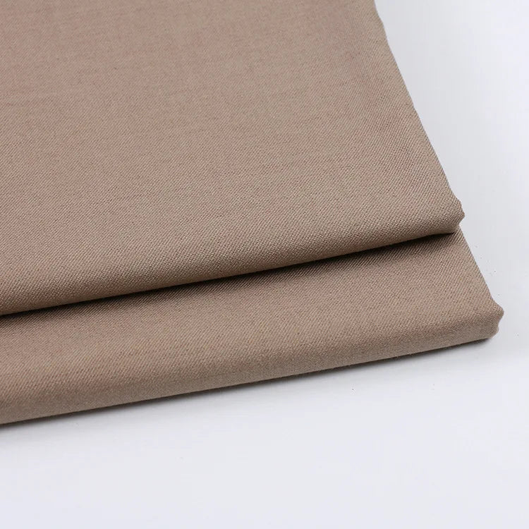Deux pièces de tissu gabardine marron clair uni au mètre de Longan Craft France sont soigneusement pliées sur une surface blanche. Cette gabardine sergée, un mélange de laine, de rayonne et de polyester, est idéale pour les vêtements décontractés.