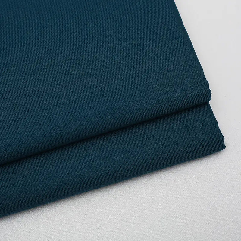 Deux pièces soigneusement pliées de tissu gabardine uni au mètre de Longan Craft France, un mélange de laine, de rayonne et de polyester de couleur sarcelle foncé, idéal pour les tenues décontractées, sont empilées sur un fond gris clair uni.