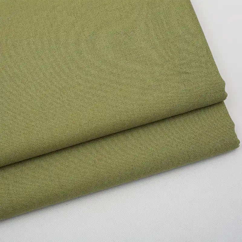Deux pièces de gabardine Tissu vert olive clair uni au mètre de Longan Craft France sont soigneusement pliées sur une surface blanche. Cette gabardine sergée, un mélange de laine, de rayonne et de polyester, est idéale pour les vêtements décontractés grâce à sa texture fine.