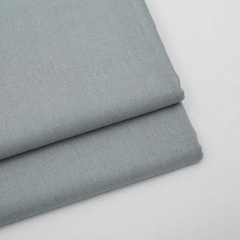 Deux pièces soigneusement pliées de Tissu gabardine gris clair uni au mètre de Longan Craft France sont empilées sur une surface blanche. Ce textile lisse et tissé serré en laine, rayonne et polyester est idéal pour les vêtements décontractés.
