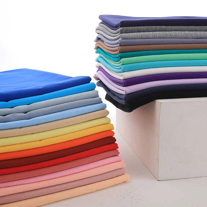 Des piles de Jersey coton élasthanne au mètre de Longan Craft France en bleu, gris, vert, violet, jaune, rouge et rose reposent sur une surface blanche. L'une d'entre elles est posée sur une boîte. Parfait tissu jersey extensible pour robes.