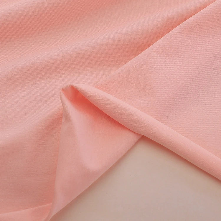 Un gros plan du Jersey coton élasthanne au mètre rose pâle de Longan Craft France, posé en douceur avec un coin plié, révèle les deux faces de ce tissu extensible parfait pour les robes de chambre.