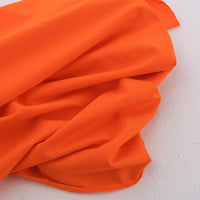 Un morceau de Jersey coton élasthanne au mètre orange vif de Longan Craft France est étalé et légèrement froissé sur une surface légère et mouchetée, parfaite pour la confection de robes et de vêtements extensibles.