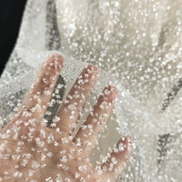 Une main touche le tissu tulle brodé de paillettes scintillantes au mètre de Longan Craft France, un tissu transparent et texturé avec des paillettes denses, idéal pour les robes de mariée et les occasions spéciales. L'arrière-plan montre davantage de tulle scintillant.