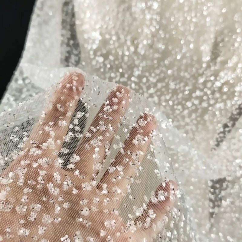 Une main touche le tissu tulle brodé de paillettes scintillantes au mètre de Longan Craft France, un tissu transparent et texturé avec des paillettes denses, idéal pour les robes de mariée et les occasions spéciales. L'arrière-plan montre davantage de tulle scintillant.