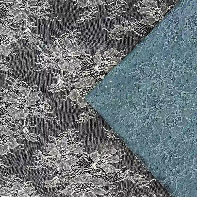 Un morceau blanc de tissu dentelle pailleté avec sequins de Longan Craft France est posé à plat, tandis qu'une variante bleu clair pliée lui est superposée. Les deux présentent des motifs floraux complexes, parfaits pour les peignoirs ou la lingerie.