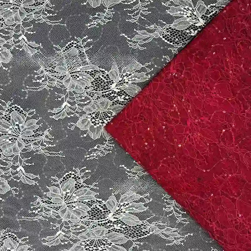 Un morceau de tissu dentelle pailleté avec sequins au mètre de Longan Craft France est posé en diagonale sur une dentelle rouge, toutes deux ornées de motifs floraux. La dentelle blanche à paillettes est transparente, tandis que le tissu rouge pour robes est opaque et texturé.