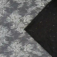 Le tissu dentelle pailleté avec paillettes au mètre de Longan Craft France est un tissu noir avec une dentelle florale blanche complexe, parfait pour les peignoirs et la lingerie. Un coin est plié pour montrer le dessous noir uni.