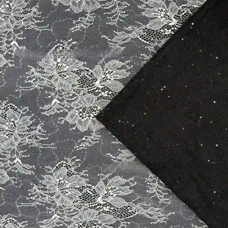 Le tissu dentelle pailleté avec paillettes au mètre de Longan Craft France est un tissu noir avec une dentelle florale blanche complexe, parfait pour les peignoirs et la lingerie. Un coin est plié pour montrer le dessous noir uni.