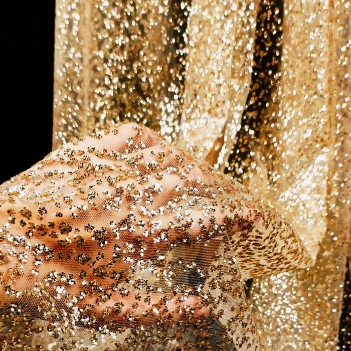 Une main tient Tissu tulle brodé de paillettes scintillantes au mètre de Longan Craft France, un tissu transparent étincelant pour robes de mariée, avec des paillettes dorées qui brillent sur un fond scintillant assorti.