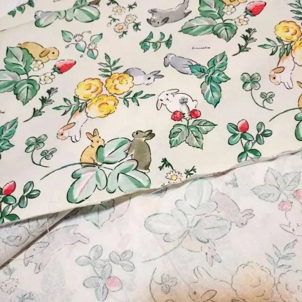 Un morceau de "Tissu motif Pâques lapin de printemps" de Longan Craft France représente des lapins, des feuilles vertes, des roses jaunes et des fraises ; un autre morceau du même tissu de coton coloré apparaît délavé en dessous.