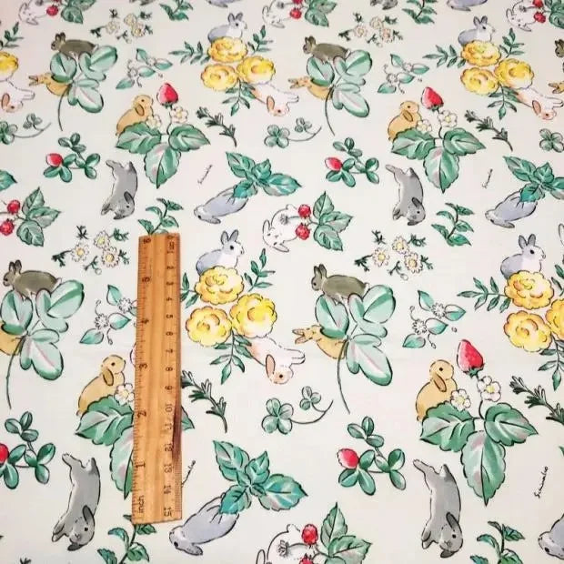 Une règle en bois repose sur le Tissu motif Pâques lapin de printemps au mètre de Longan Craft France, qui présente des lapins, des fleurs, des feuilles et des fraises illustrés sur un tissu de coton léger - idéal pour les créations et décorations printanières.