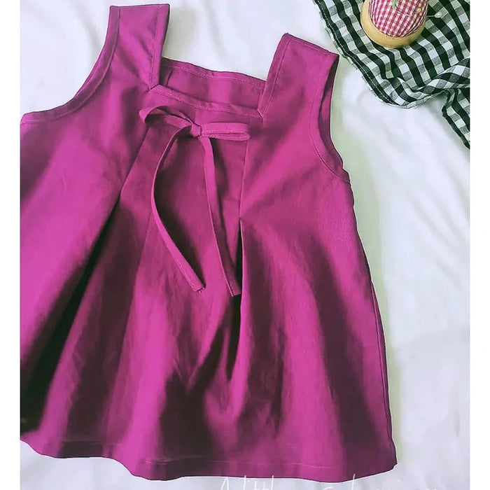 Le "Patron de couture gratuit pour blouse sans manches à encolure carrée" de Longan Craft France est présenté sur une surface blanche à côté d'un tissu à carreaux noirs et blancs et d'un petit récipient à couvercle rose.