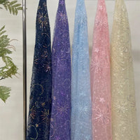 Cinq tulles transparents de Longan Craft France sont suspendus verticalement dans des couleurs bleu foncé, violet, bleu clair, rose et beige. Chaque "Tulle brodé de sequins étoiles et lunes" est orné d'étoiles brodées et de feux d'artifice. Un peu de verdure apparaît sur la gauche.