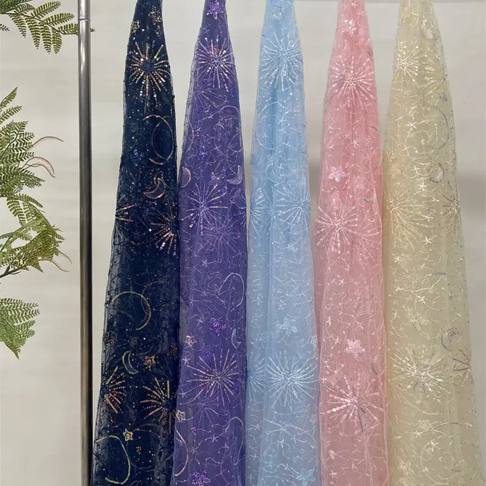 Cinq tulles transparents de Longan Craft France sont suspendus verticalement dans des couleurs bleu foncé, violet, bleu clair, rose et beige. Chaque "Tulle brodé de sequins étoiles et lunes" est orné d'étoiles brodées et de feux d'artifice. Un peu de verdure apparaît sur la gauche.