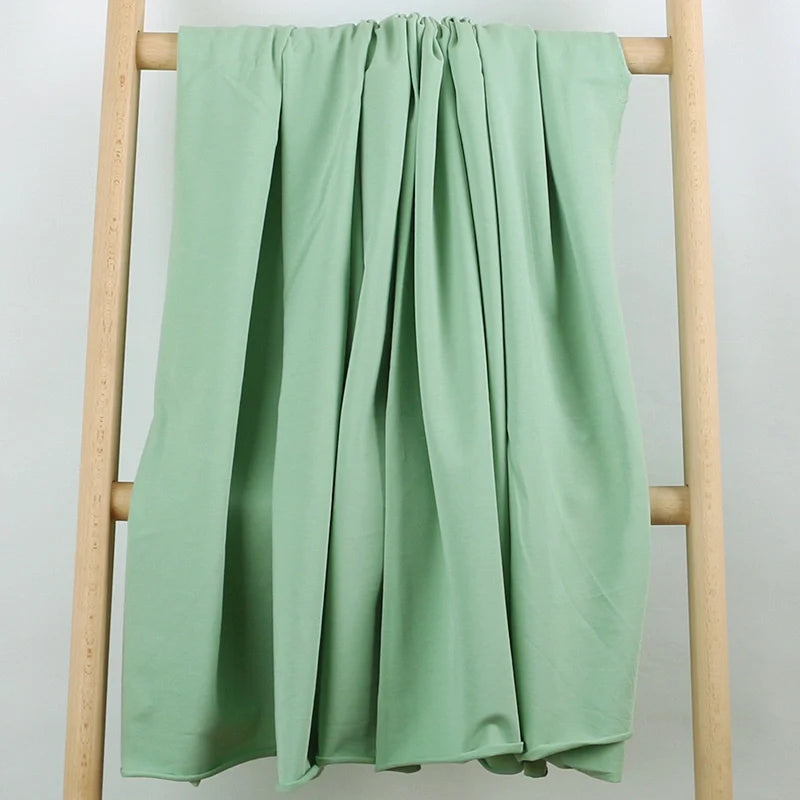 Un Tissu French Terry en coton extensible vert clair de Longan Craft France est suspendu en plis amples et verticaux au-dessus d'une échelle en bois sur un fond blanc. Ce tissu confortable et extensible est idéal pour les vêtements de sport et est vendu au mètre.