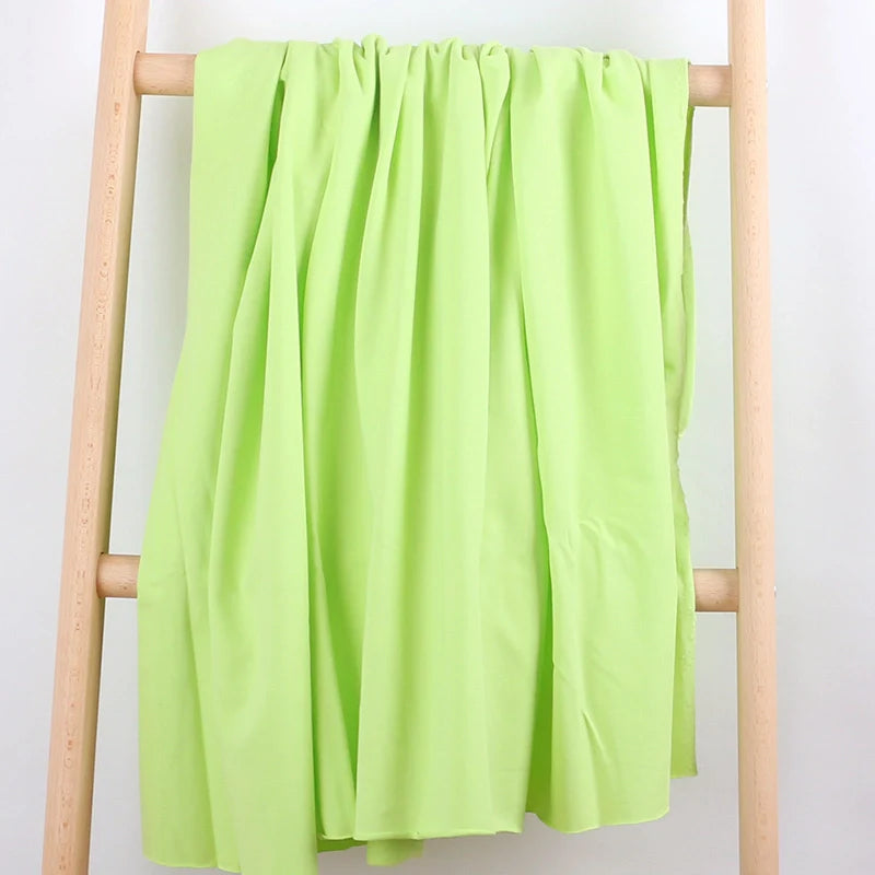 A Tissu French Terry en coton extensible au mètre de Longan Craft France, dans une teinte vert clair, est drapé sur une échelle en bois clair devant un fond blanc, formant des plis souples parfaits pour la confection de vêtements de sport.