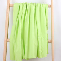 A Tissu French Terry en coton extensible au mètre de Longan Craft France, dans une teinte vert clair, est drapé sur une échelle en bois clair devant un fond blanc, formant des plis souples parfaits pour la confection de vêtements de sport.