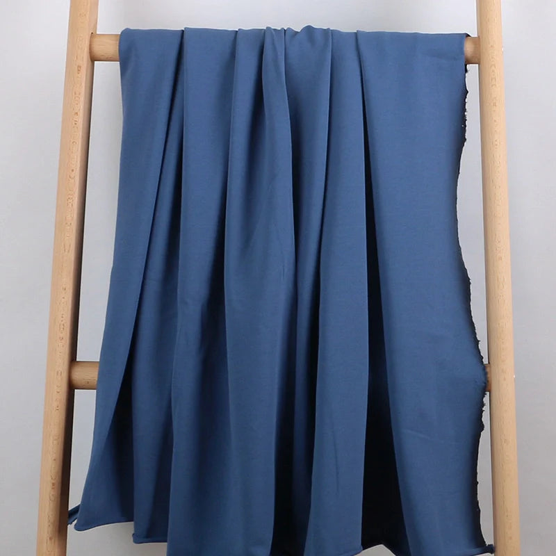 Un morceau de Tissu French Terry en coton extensible de Longan Craft France en bleu moyen, idéal pour les vêtements de sport, est drapé sur une échelle en bois clair avec un bord subtilement inachevé ; le fond est uni et de couleur claire.