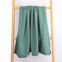 Un pli en tissu éponge en coton extensible de Longan Craft France, vert sauge, est drapé sur une échelle en bois clair devant un mur blanc texturé - confortable et idéal pour les vêtements de sport, vendu au mètre.