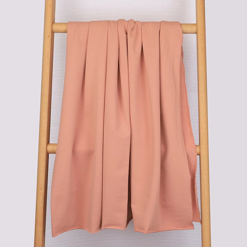 Un tissu de coton French Terry multicolore et extensible de Longan Craft France est drapé sur une échelle en bois contre un mur clair. Ce tissu confortable et tricoté est parfait pour les vêtements de sport et ajoute une touche douillette à la scène.