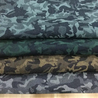 Quatre pièces pliées de Tissu denim jacquard camouflage extensible au mètre de Longon Craft France sont empilées, chacune avec un motif de camouflage coloré distinct dans les tons bleu-gris, vert, marron et gris, montrées de près pour faire ressortir les détails.