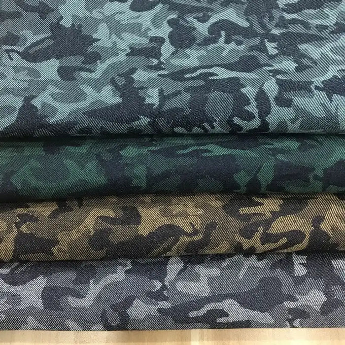 Quatre pièces pliées de Tissu denim jacquard camouflage extensible au mètre de Longon Craft France sont empilées, chacune avec un motif de camouflage coloré distinct dans les tons bleu-gris, vert, marron et gris, montrées de près pour faire ressortir les détails.