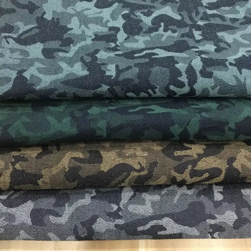 Quatre pièces pliées de Tissu denim jacquard camouflage extensible au mètre de Longon Craft France sont empilées, chacune avec un motif de camouflage coloré distinct dans les tons bleu-gris, vert, marron et gris, montrées de près pour faire ressortir les détails.