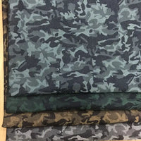 Trois pièces de Tissu denim jacquard camouflage extensible au mètre de Longon Craft France - bleu-gris, vert et marron - aux motifs camouflage colorés sont empilées sur une surface claire.