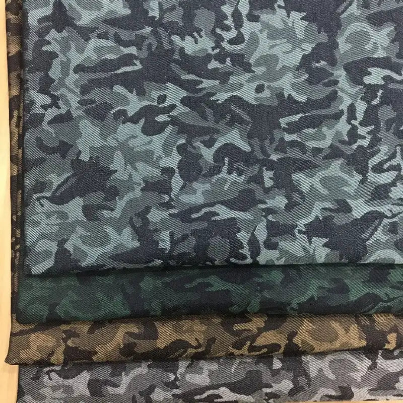 Trois pièces de Tissu denim jacquard camouflage extensible au mètre de Longon Craft France - bleu-gris, vert et marron - aux motifs camouflage colorés sont empilées sur une surface claire.